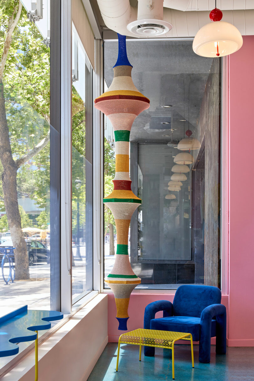 A colorful sculptural installation hangs in front of a large window next to a blue chair, yellow table, and pink wall in a modern, brightly lit interior.
