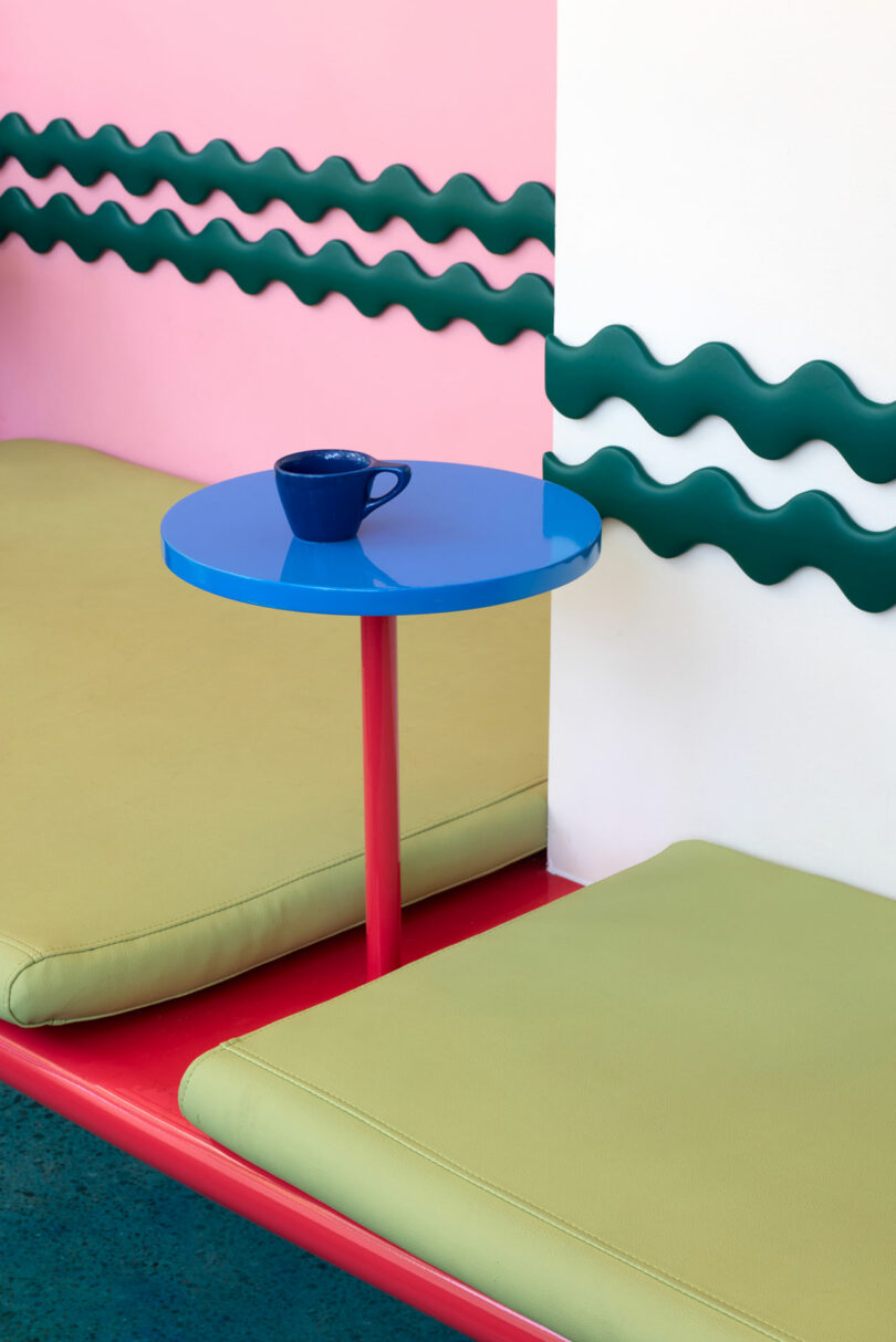 A small blue cup sits on a round blue table between two light green cushions against a pink and white wall with green wavy trim.