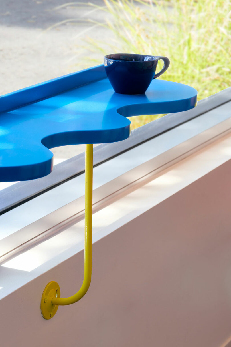 A blue, irregularly-shaped wall-mounted shelf with a yellow support holds a dark blue cup, positioned by a window with sunlight and greenery visible outside.