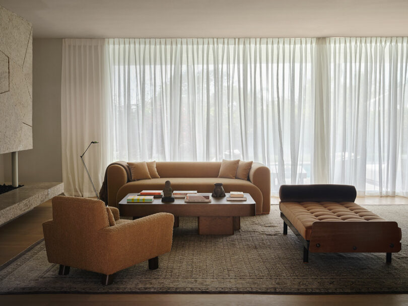 A modern OSKLO-inspired living room with a tan sofa, matching armchair, and chaise lounge around a low coffee table on a patterned rug; sheer curtains cover the large window in the background.