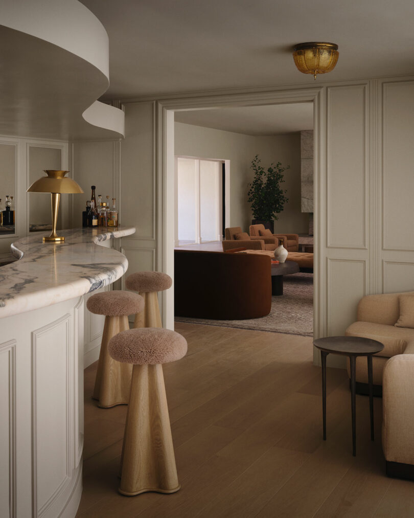 Modern home interior by OSKLO featuring a curved marble bar with gold accents, plush barstools, neutral-toned seating area, and warm lighting.