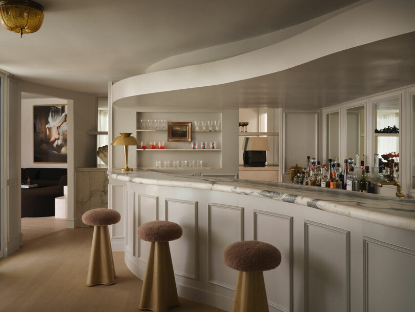 Modern bar interior by OSKLO with a curved marble counter, beige paneled front, plush round stools, glass shelves with bottles and glasses, and soft ambient lighting.
