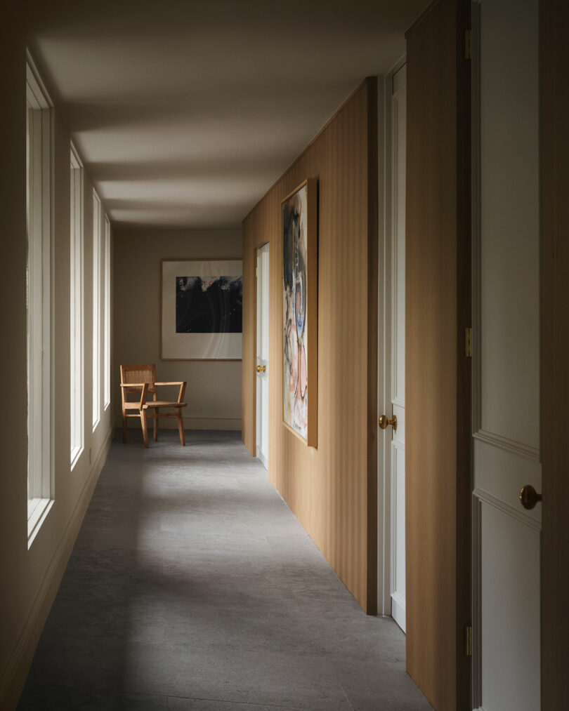 A hallway with large windows on one side, wooden paneled walls, white doors, and a wooden chair beside framed artwork at the far end reflects OSKLO’s signature blend of warmth and modernity.