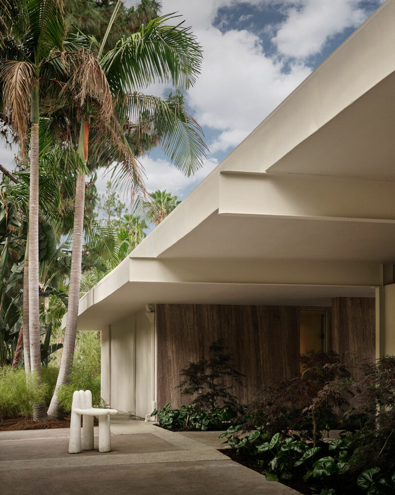 Modern house exterior with flat roof, beige walls, and tropical landscaping by OSKLO, featuring a white sculptural bench under tall palm trees on a cloudy day.