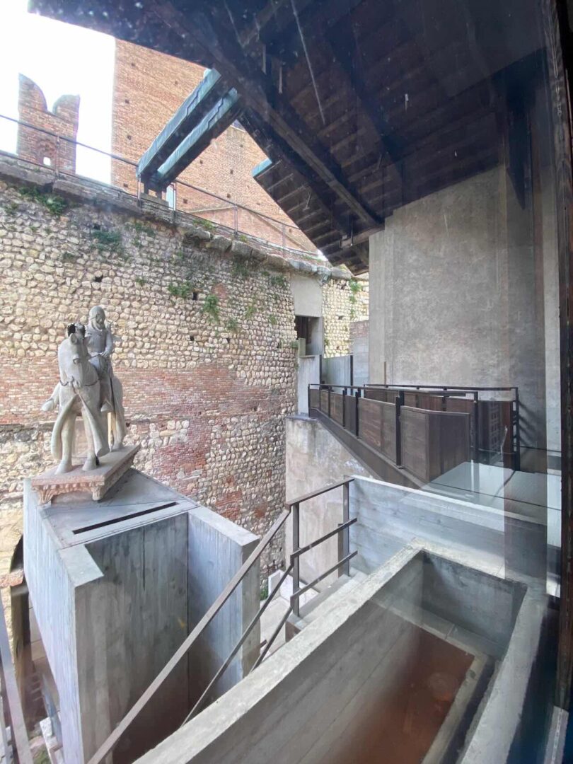Stone statue of two figures on a concrete platform, surrounded by stone walls and modern architectural elements, seen through a glass window—an ideal spot to Take 5 and appreciate art and architecture.
