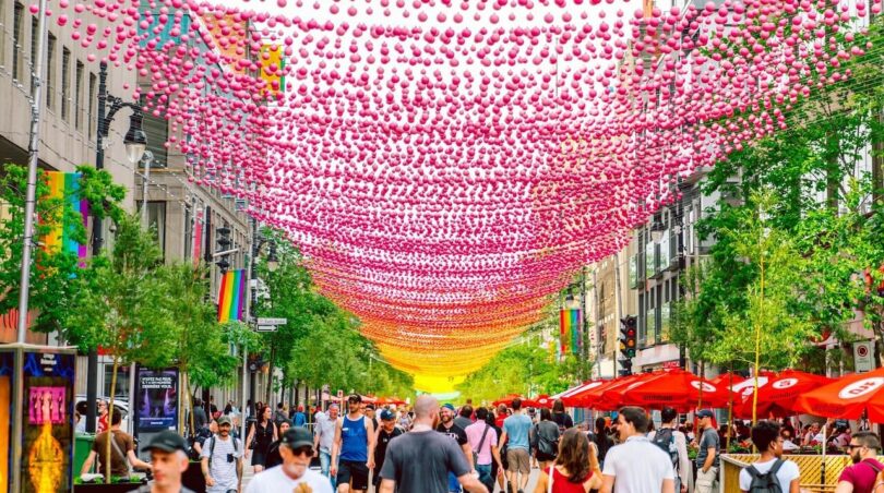Crowds walk along a city street decorated with pink and yellow hanging spheres, with trees, red umbrellas, and buildings on either side.
