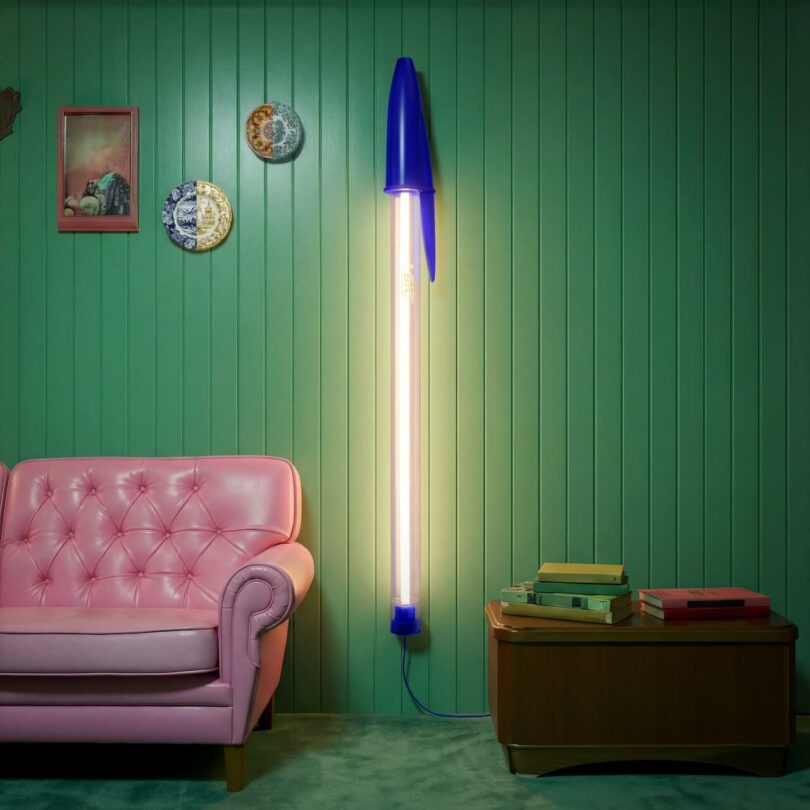 A tall floor lamp designed to look like a blue pen stands next to a pink couch, green paneled wall, and a wooden side table with books and plates as decor.