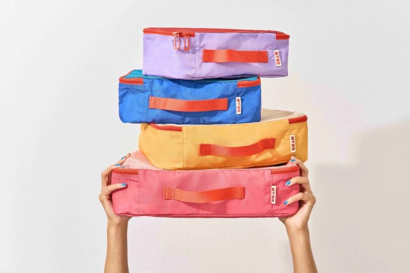 Four colorful packing cubes stacked on top of each other with two hands at the bottom of the photo holding them up. From top to bottom, there's a (small) purple packing cube, then (small) blue, (medium) orange, and (large) red - all with red handles and zipper details.