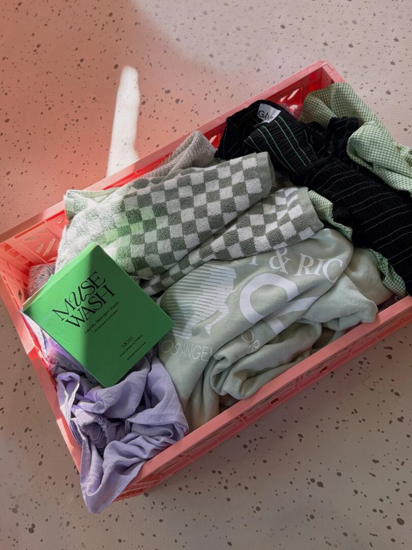 A pink crate holds an assortment of clothes with MUSEWASH's green detergent sheet box laying on top of it