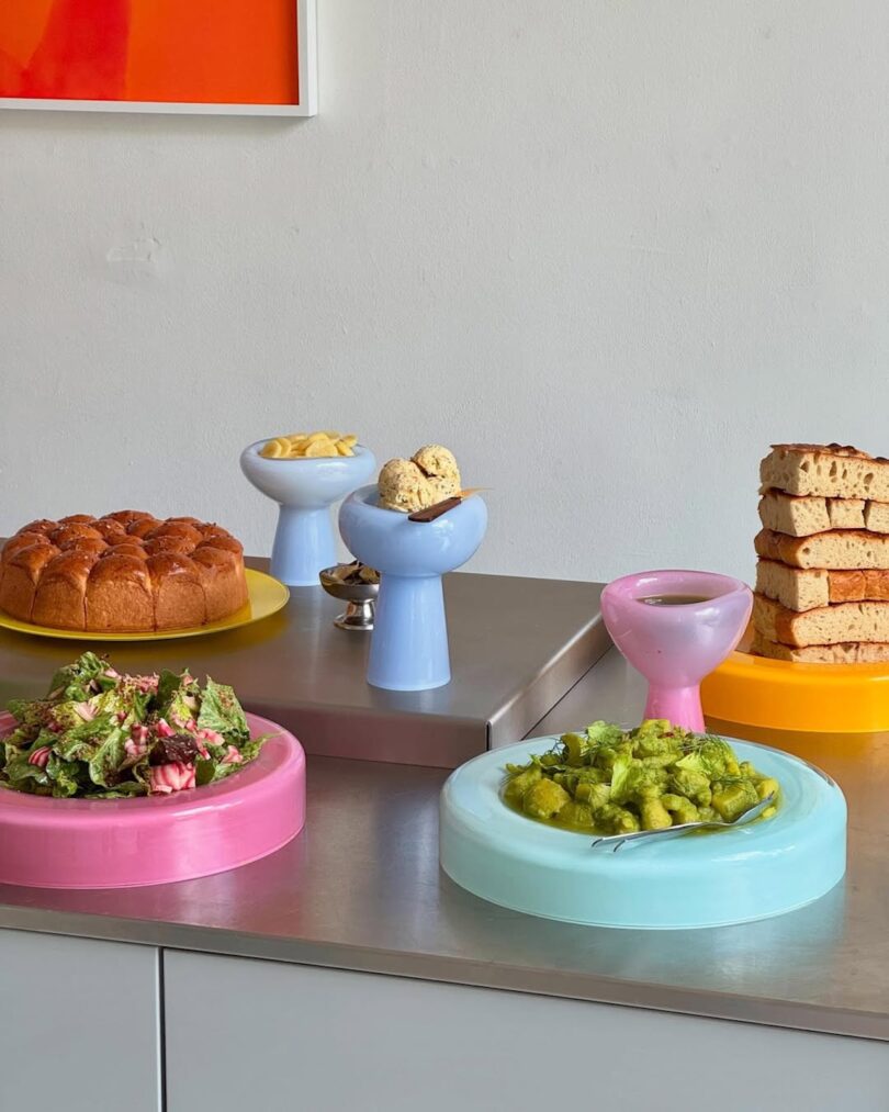 A table with assorted foods on colorful pastel serving glassware, including salad, bread, cake, cookies, chips, and a green pasta dish.
