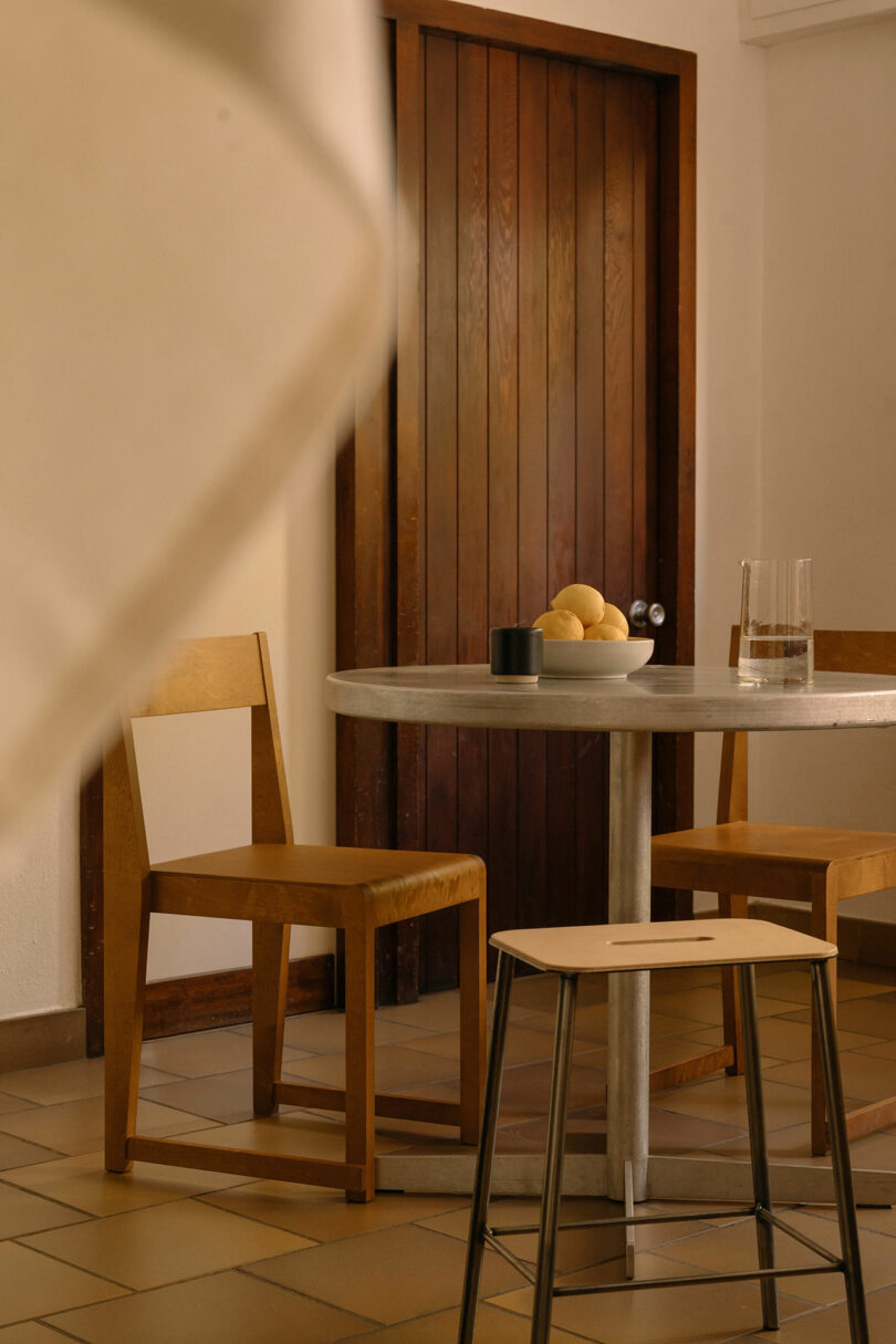A Tasca Table FRAMA sits between two wooden chairs, topped with a bowl of lemons, a glass of water, and a cup in a room featuring tiled floors and a wooden door.
