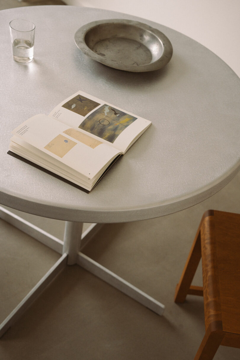 A Tasca Table FRAMA holds an open art book, a glass of water, a metal bowl, and is paired with a wooden chair beside it.