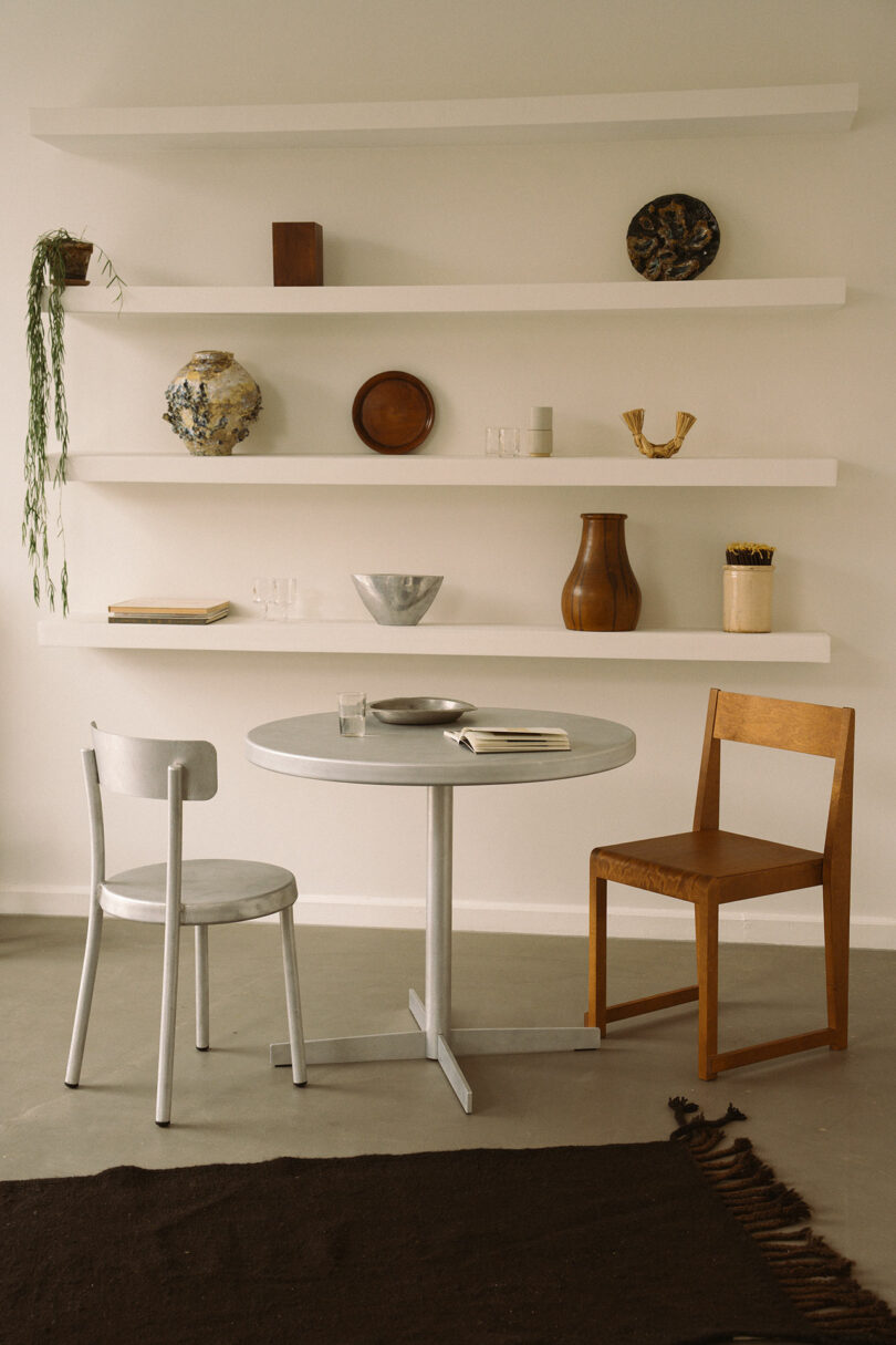 A round Tasca Table FRAMA with two chairs, an open book, and a glass sits in front of white shelves displaying decorative objects, vases, and bowls against a neutral wall.