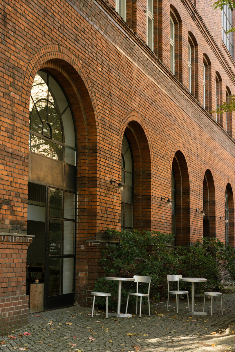 Outdoor tables and chairs, including the Tasca Table FRAMA, are arranged on a cobblestone patio next to a brick building with tall arched windows and some greenery.