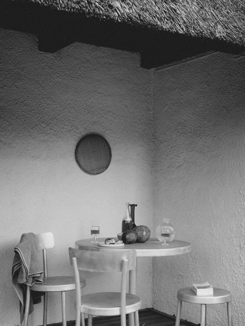 Black and white photo of a small round Tasca Table FRAMA with two chairs on a patio, holding a pitcher, two glasses, a round fruit, and a book; a woven decor hangs on the wall.