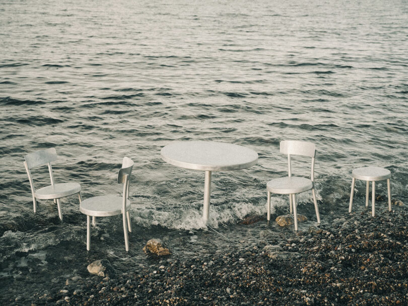 Four metal chairs and a Tasca Table from FRAMA are partially submerged at the edge of a rocky shoreline, with waves from the water reaching the furniture.