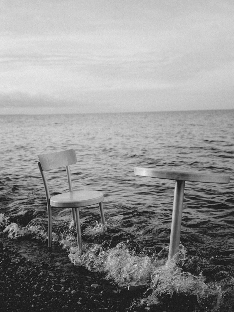 A wooden chair and a Tasca Table FRAMA stand partially submerged in shallow water at the edge of a rocky shore.