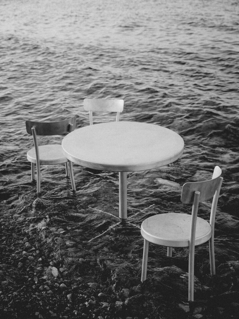 A Tasca Table FRAMA with three chairs stands in shallow water at the edge of a rocky shoreline, gentle waves surrounding the legs. Black and white image.