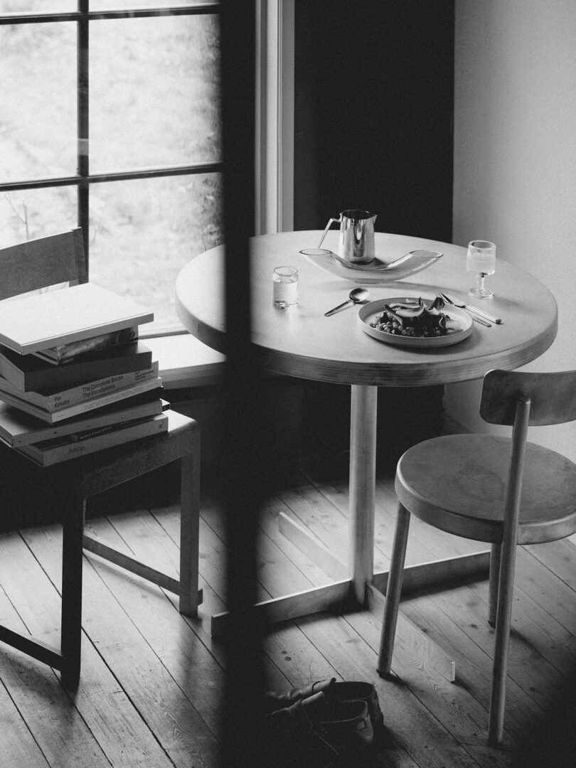 A Tasca Table FRAMA stands round with a plate of food, utensils, a glass of water, and a teapot beside two chairs—one near a stack of books by the window. Black and white photo.