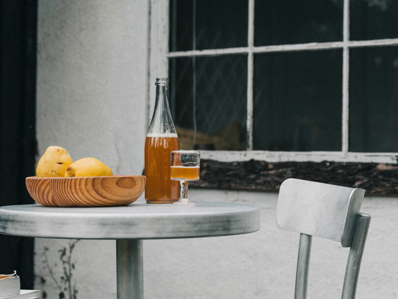 A Tasca Table from FRAMA holds a wooden bowl of pears, a glass bottle of amber liquid, and a filled glass, placed beside a metal chair and a window.