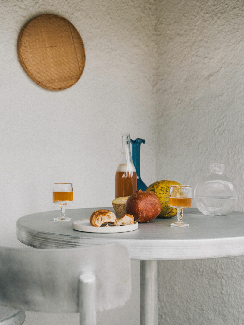 A small round Tasca Table from FRAMA with two glasses of amber liquid, a bottle, a glass pitcher, a plate with pastries, and assorted fruits set against a textured white wall.