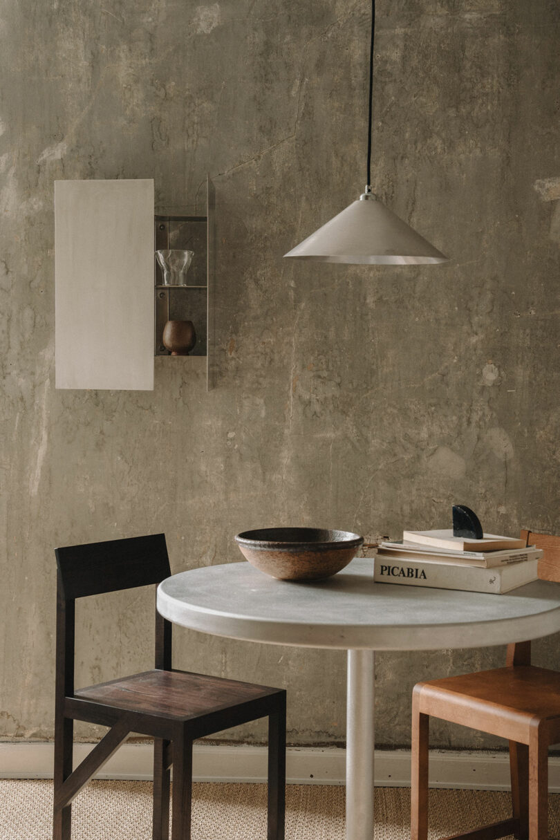 A Tasca Table from FRAMA sits between two chairs, topped with a wooden bowl and books, against a textured concrete wall beneath a hanging pendant light.