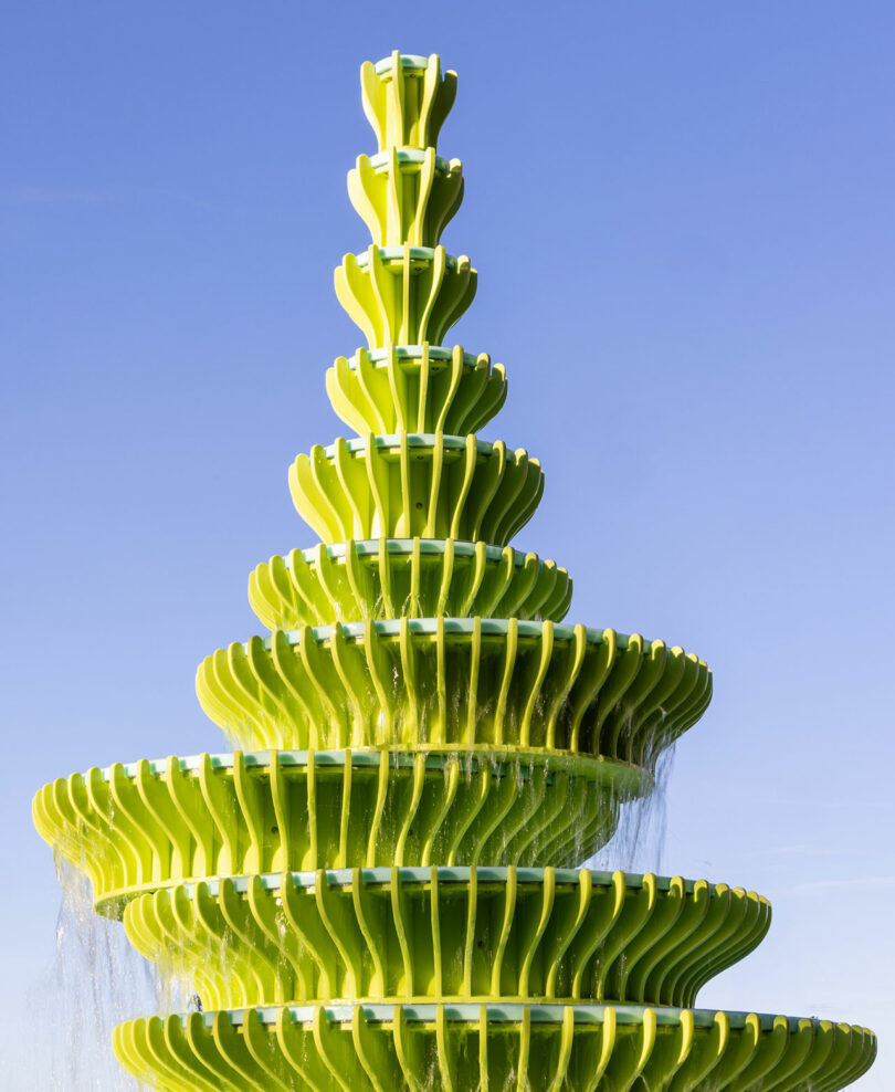 A modern, tiered bright green fountain with cascading water against a clear blue sky.