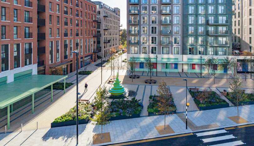 Aerial view of a modern urban square with landscaped gardens, benches, a green-striped sculpture, and surrounding apartment buildings. One person walks along the paved path.