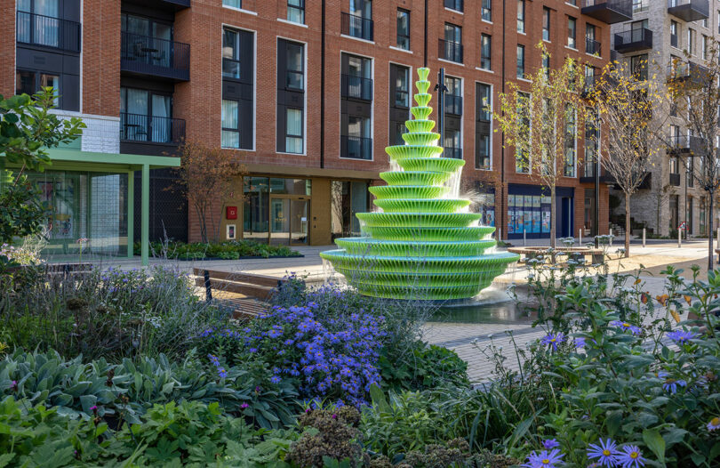 A tiered, green modern fountain stands in a landscaped urban courtyard with purple flowers and plants, surrounded by apartment buildings.