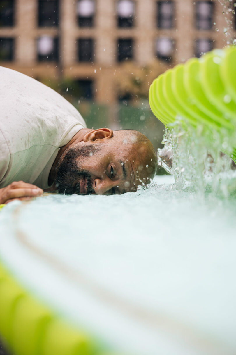 A man rests his head sideways on the edge of a water fountain, with water splashing nearby and a blurred building in the background.