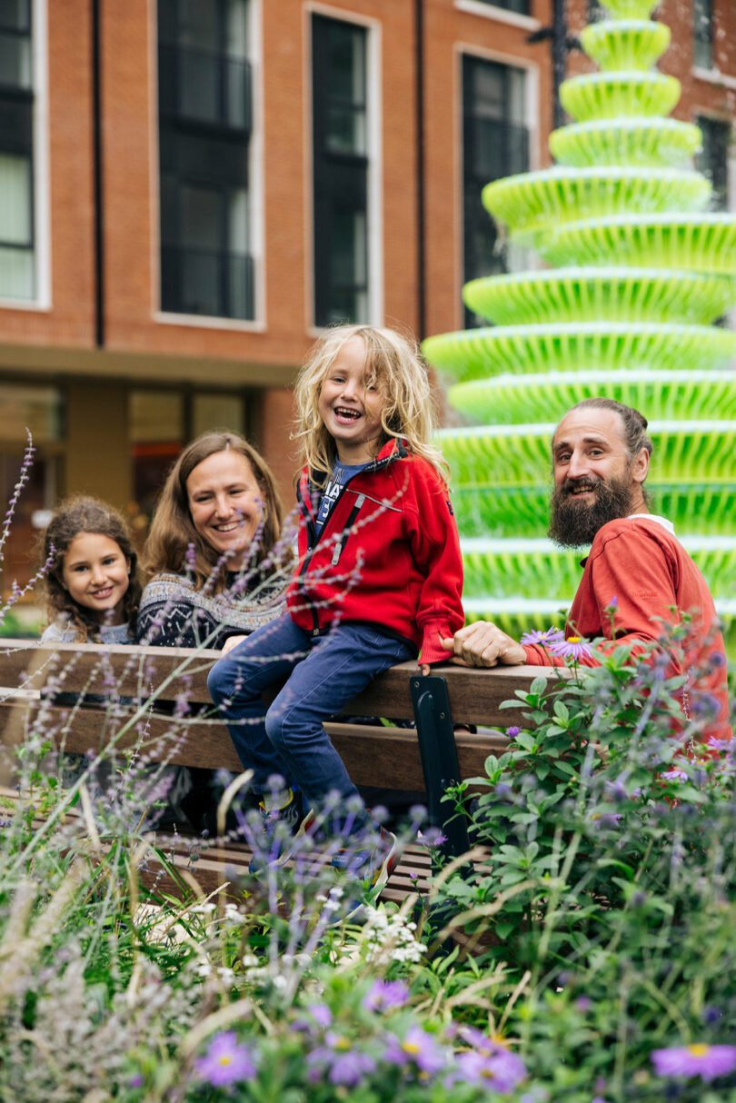 A family of four sits and stands around a park bench, smiling, with flowers in the foreground and a bright green fountain in the background.