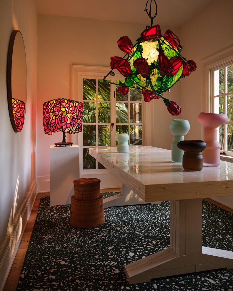 A modern dining room curated by The Future Perfect, featuring a white table, pastel vases, a wooden stool, a floral-patterned lamp and chandelier, and large windows with a garden view.