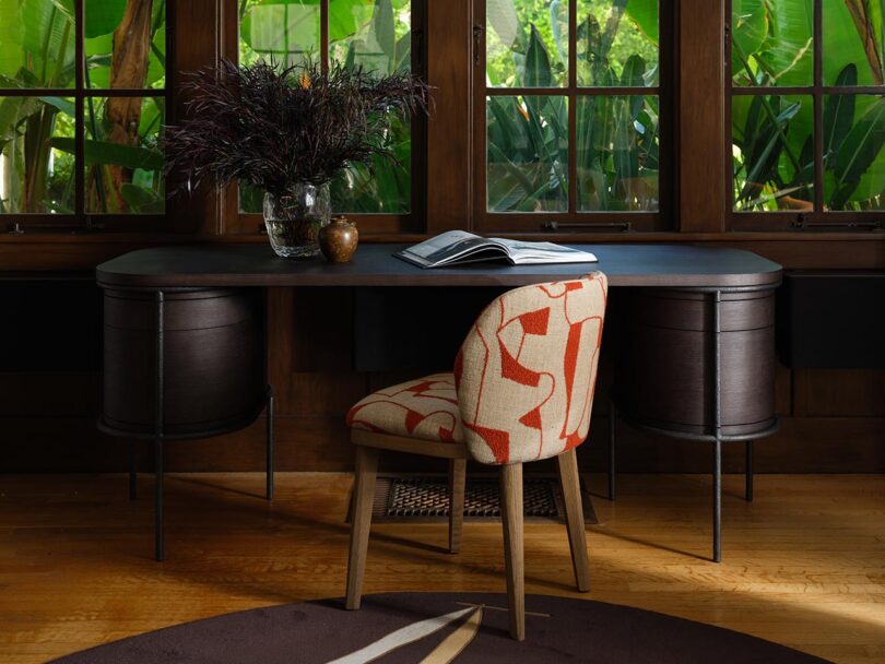 A wooden desk from The Future Perfect with an open book, a vase of dark foliage, and a small brown jar sits in front of large windows; a patterned chair is positioned in front of the desk.