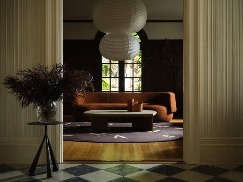 A modern living room curated by The Future Perfect, featuring a curved brown sofa, round paper lanterns, a rug with leaf motifs, and a side table topped with a glass vase of dark foliage.