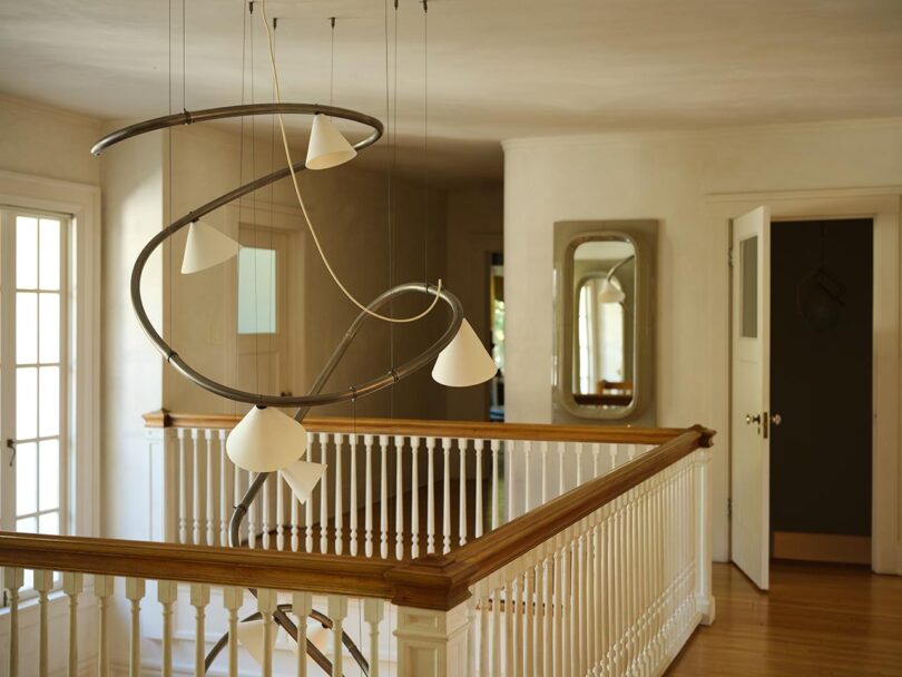 Modern chandelier from The Future Perfect with spiral metal frame and white shades hangs over a wooden railing in a bright hallway with doors, windows, and a wall mirror.