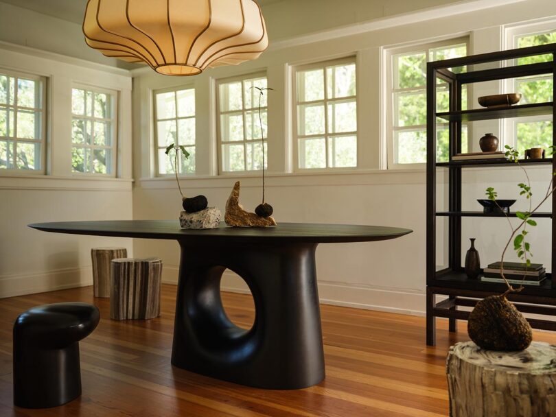 Modern dining room with a sculptural black table from The Future Perfect, small black stool, tree stump side tables, open shelving, and large pendant light; sunlight streams through multiple windows.