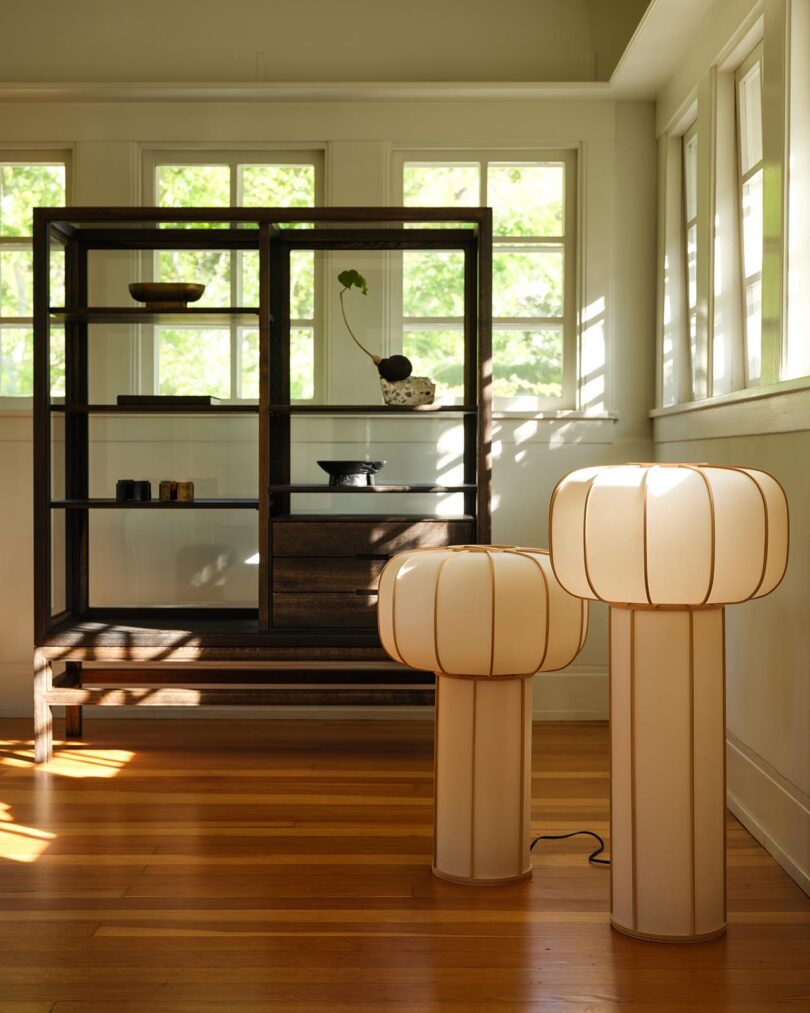 A minimalist room curated by The Future Perfect, featuring wooden floors, a dark open shelving unit with ceramics, and two tall, glowing white lamps with rounded shades. Sunlight streams in through large windows.