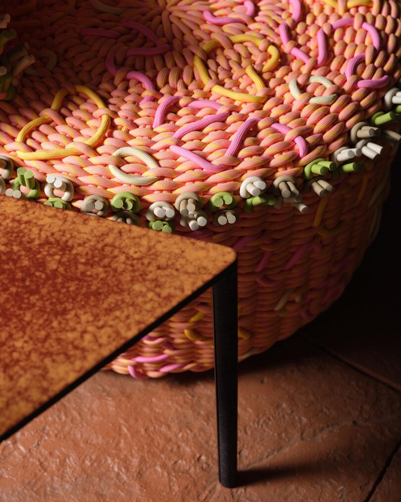 A close-up of a woven chair from The Future Perfect, featuring colorful, intertwined strands in pink, yellow, green, and orange beside a rust-colored table on a textured floor.