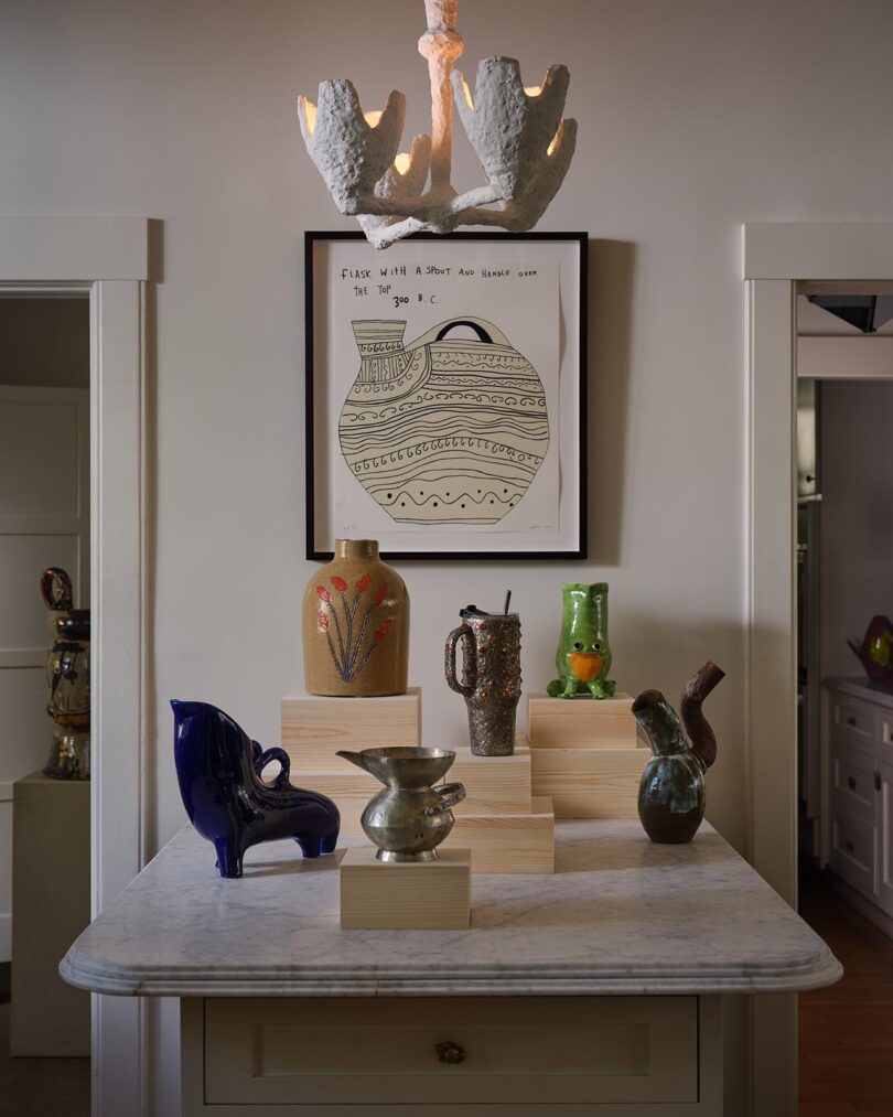 A marble-topped table from The Future Perfect displays various ceramic vases and pitchers on wooden blocks, set beneath a framed drawing and a white sculptural chandelier in a refined home interior.