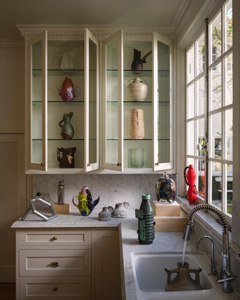 A kitchen corner with glass-front cabinets displaying pottery and vases from The Future Perfect, a marble countertop, decorative objects, and a large window above the sink.
