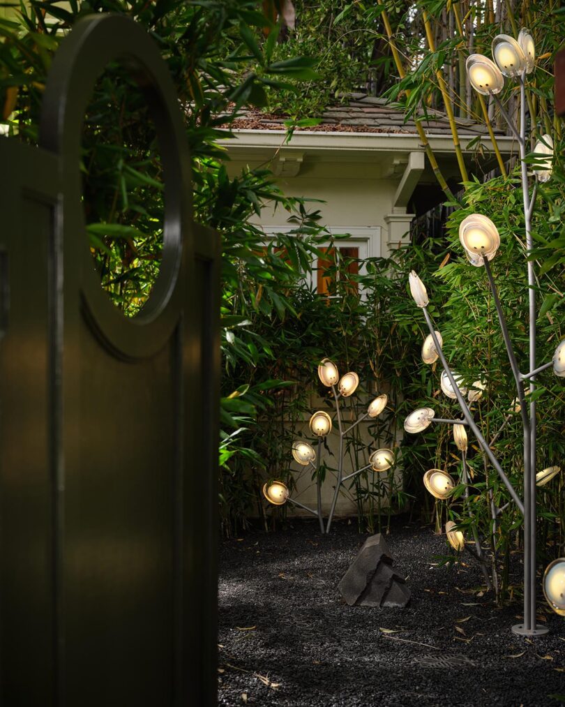 A garden with gravel ground features clusters of modern outdoor lights resembling flowers, curated by The Future Perfect, surrounded by lush green plants near a white house.