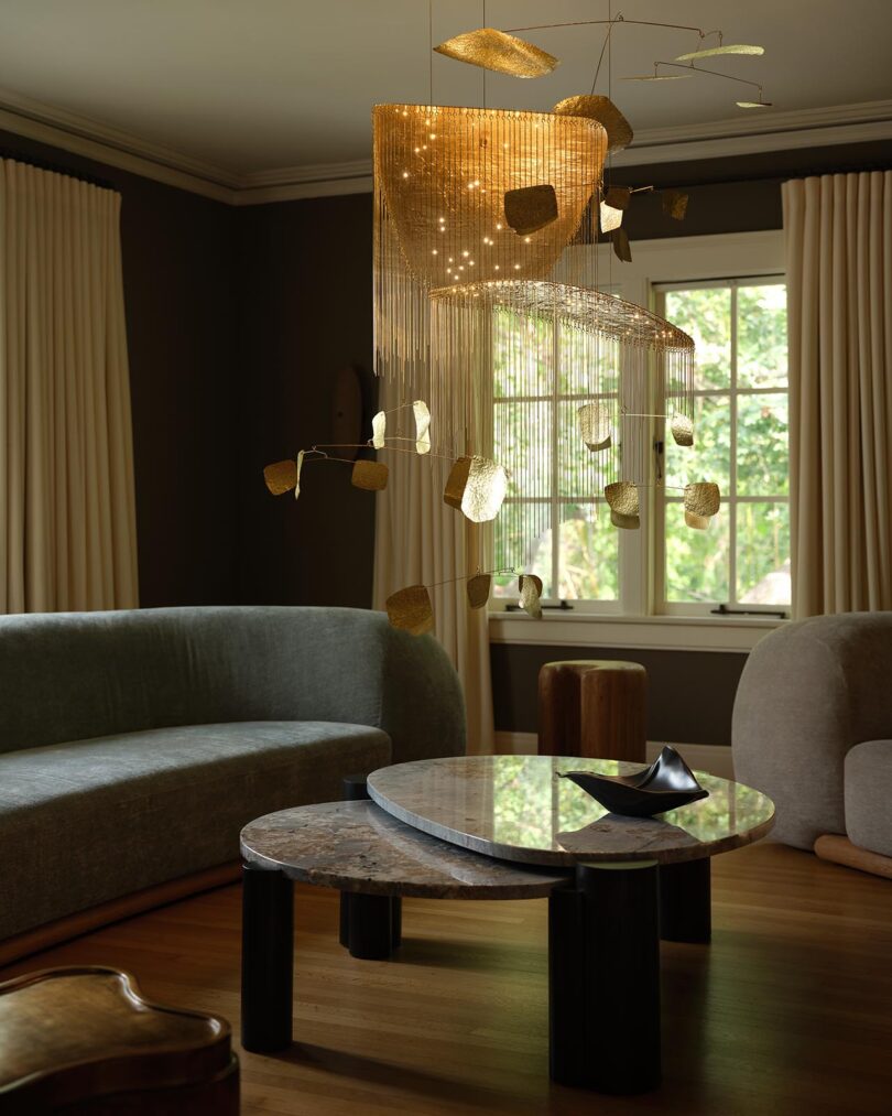 Modern living room curated by The Future Perfect, featuring curved sofas, a marble coffee table, sculptural decor, and a gold leaf-like chandelier; large window with cream curtains in the background.