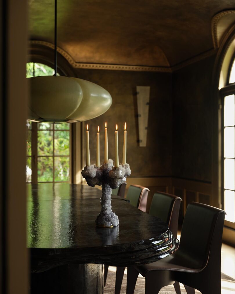 A dining room with a dark, glossy table, modern chairs from The Future Perfect, a cluster candle holder with five lit candles, large windows, and a rounded pendant light overhead.
