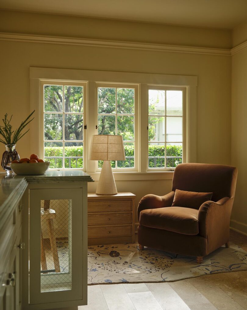 A cozy reading nook with a brown armchair, side table with a lamp, and a window overlooking a green garden from The Future Perfect.