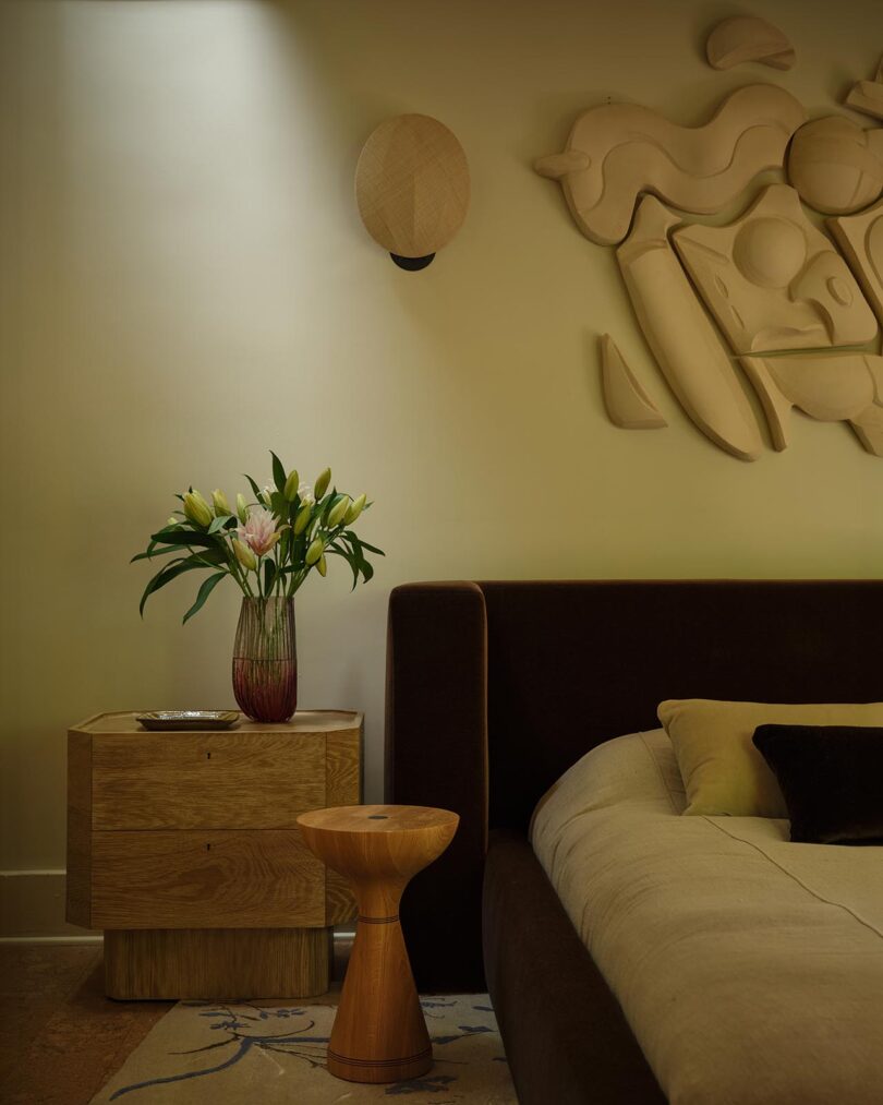 A modern bedroom from The Future Perfect features a wooden nightstand with a vase of lilies, a low bed with neutral bedding, a small wooden stool, and abstract wall art above the bed.