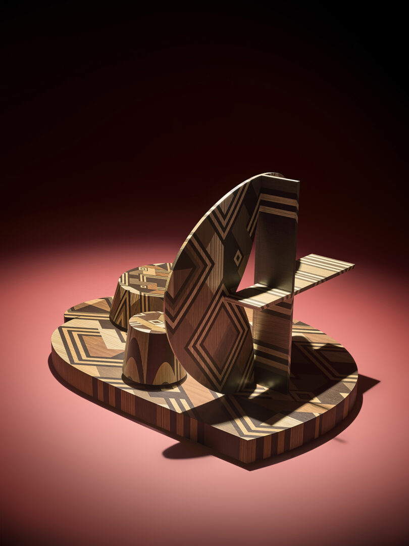 Geometric abstract wooden sculpture with patterned surfaces sits on a red background under a spotlight.