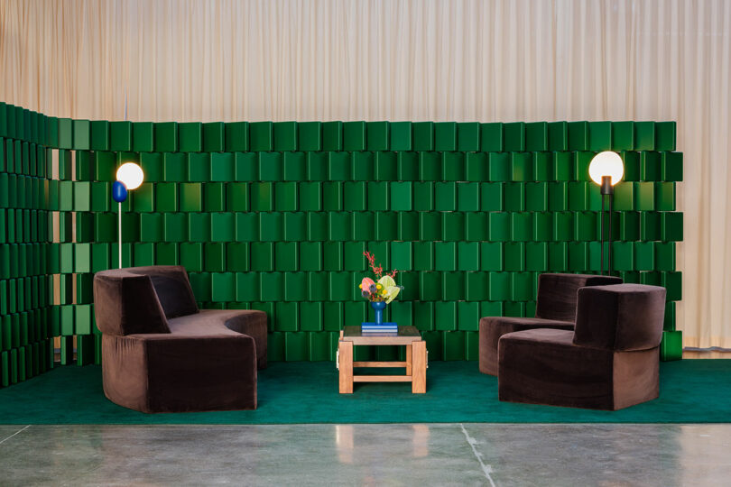 Two dark brown lounge chairs face each other with a small wooden table and a vase of flowers between them, set against a green paneled wall and green carpet, with two round wall lamps.