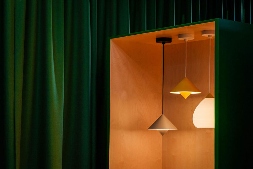 Three pendant lights with different geometric shades hang inside a wooden display box, set against a background of dark green curtains.