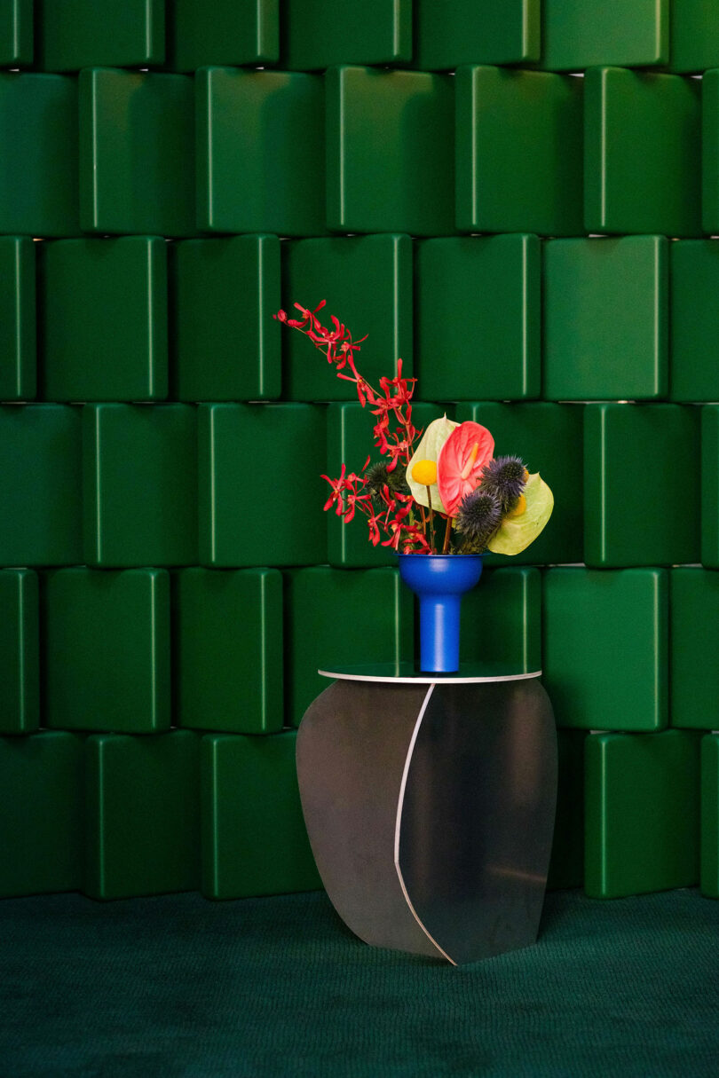 A blue vase with colorful flowers sits on a curved metallic table in front of a textured green wall.