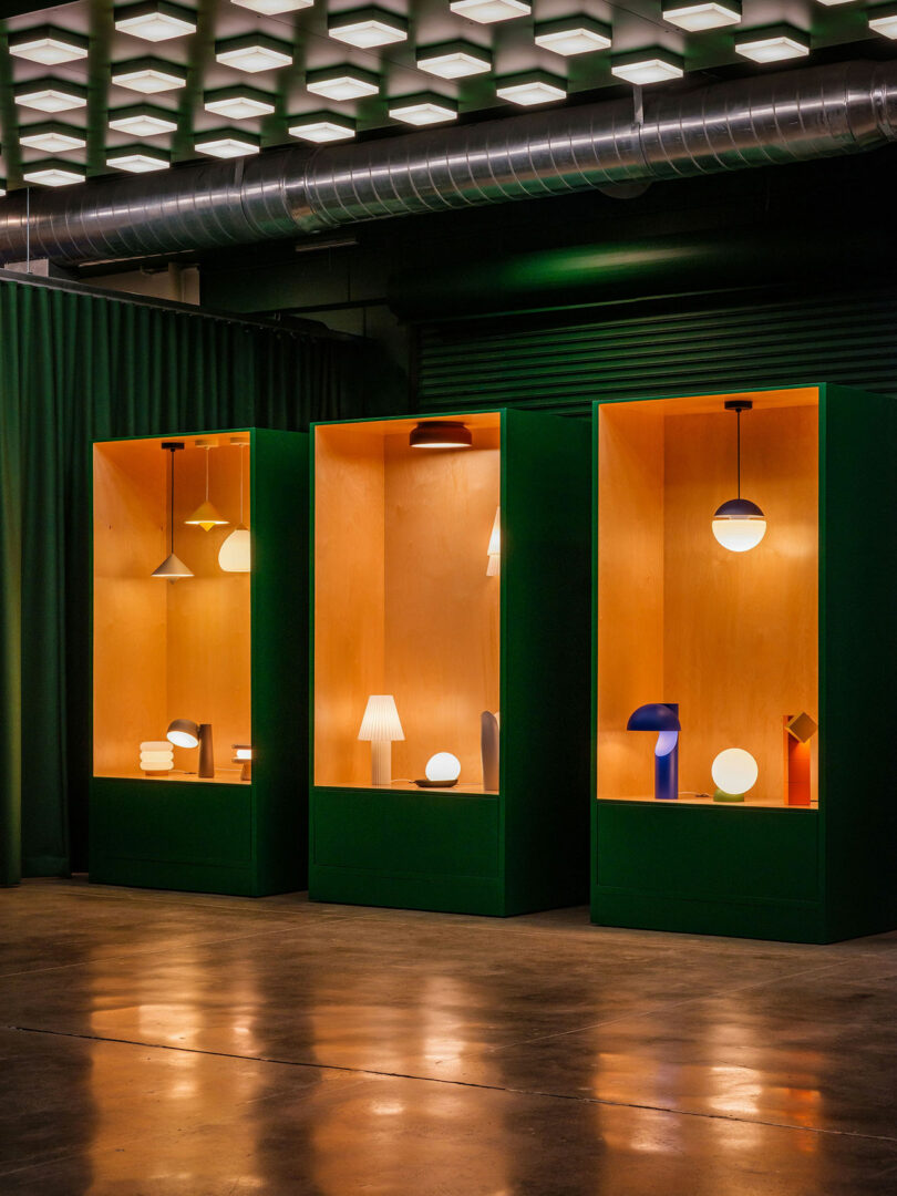 Three green display cases with wooden interiors showcase various modern table and pendant lamps, illuminated against an industrial setting with exposed ductwork and ceiling lights.